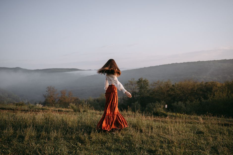 A woman in a flowing skirt walks through a misty meadow with hills in the background.
