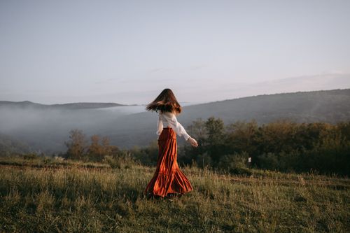 Free A woman in a flowing skirt walks through a misty meadow with hills in the background. Stock Photo