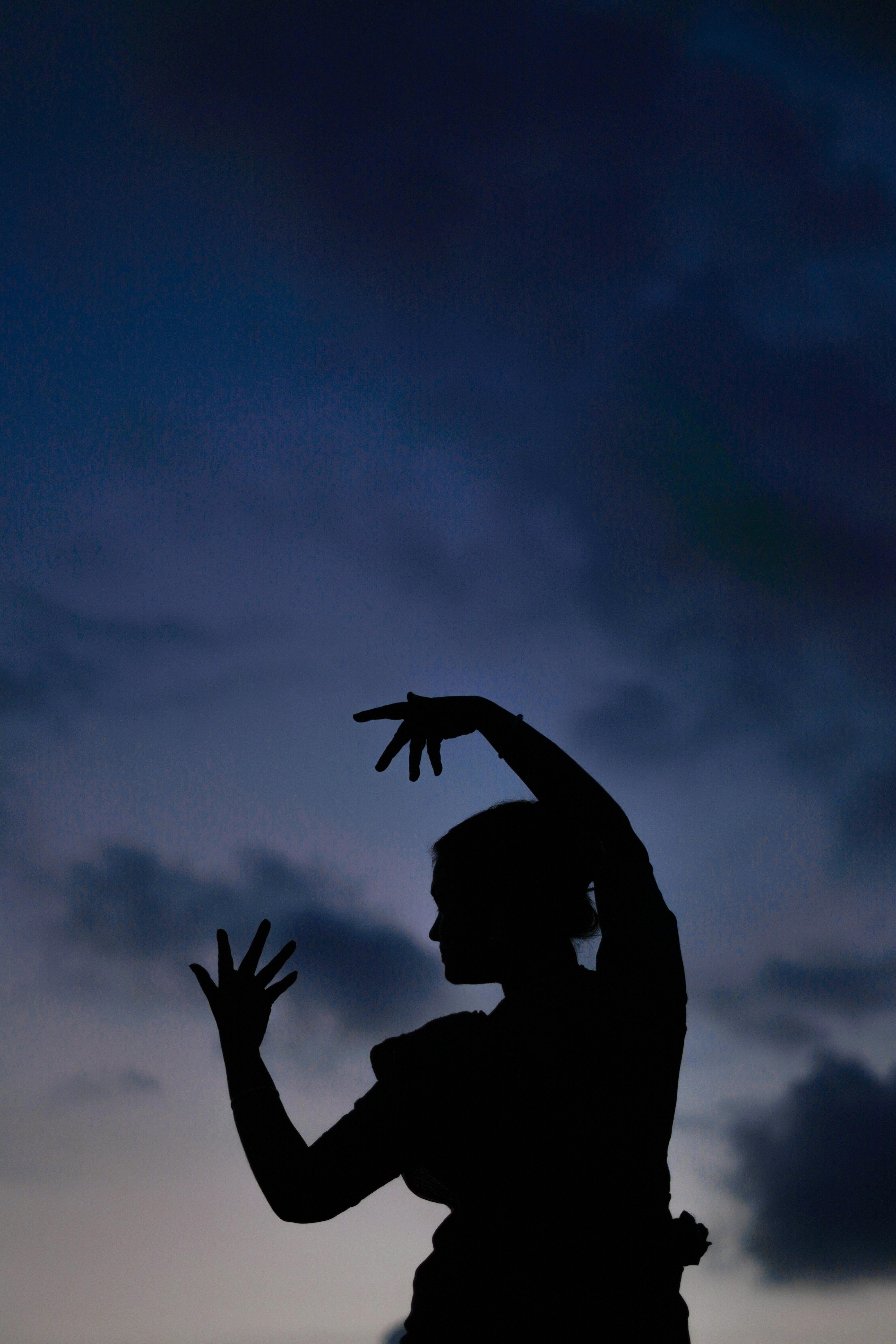 Silhouette of a woman performing traditional dance with arms raised against a sunset sky.