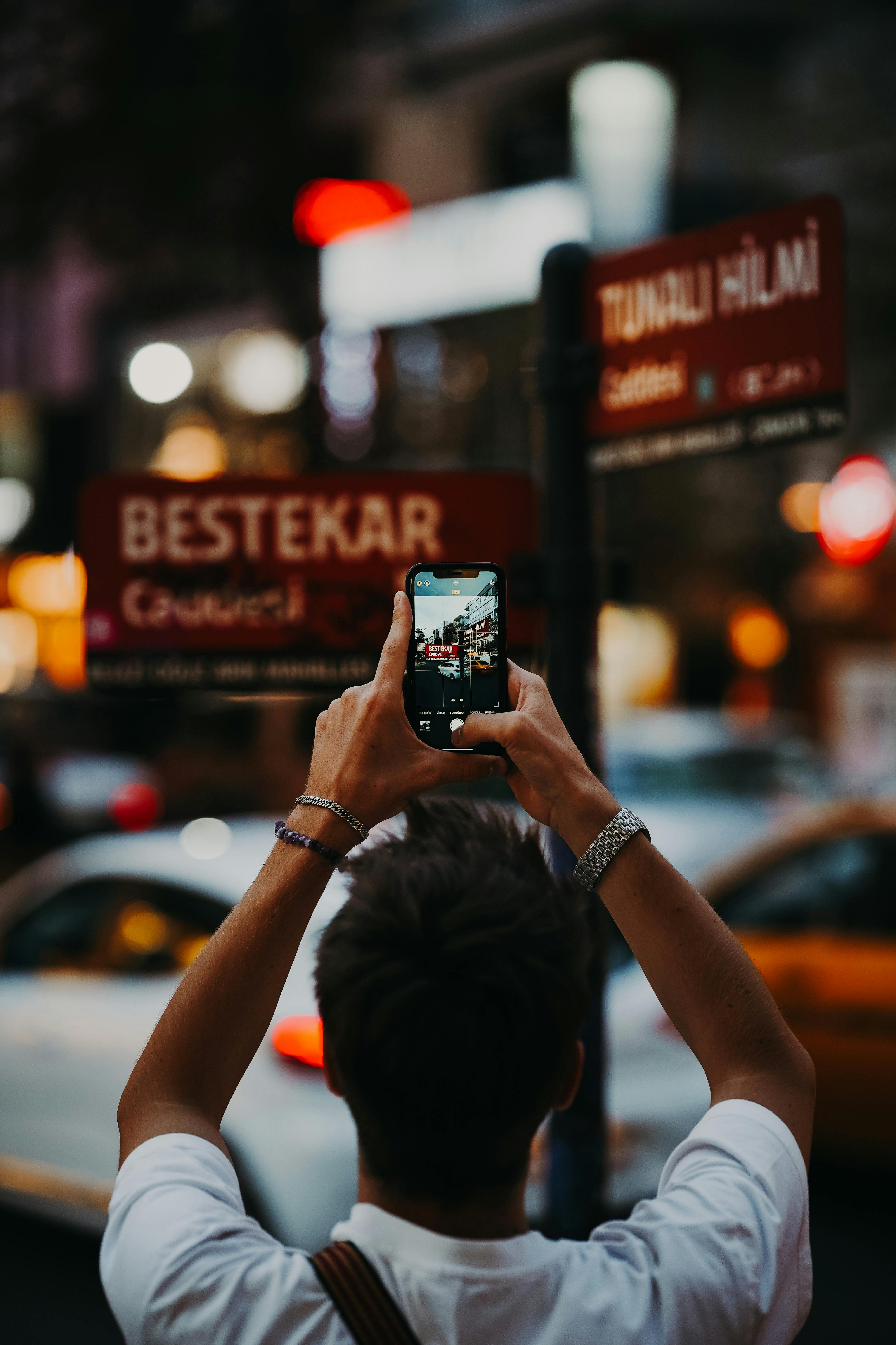 Back view of a man capturing urban Ankara streets with his smartphone at night.