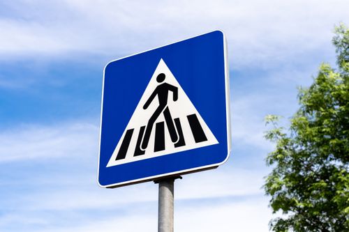 Free A road sign indicating a pedestrian crossing, set against a clear blue sky with clouds and a tree in view. Stock Photo
