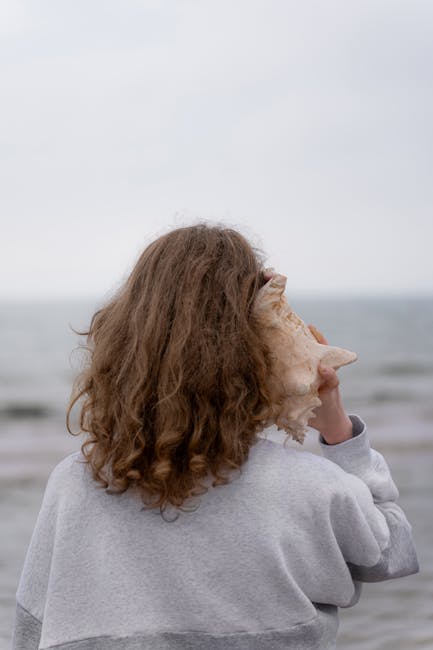 A young woman listens to a large seashell on a cloudy beach day.
