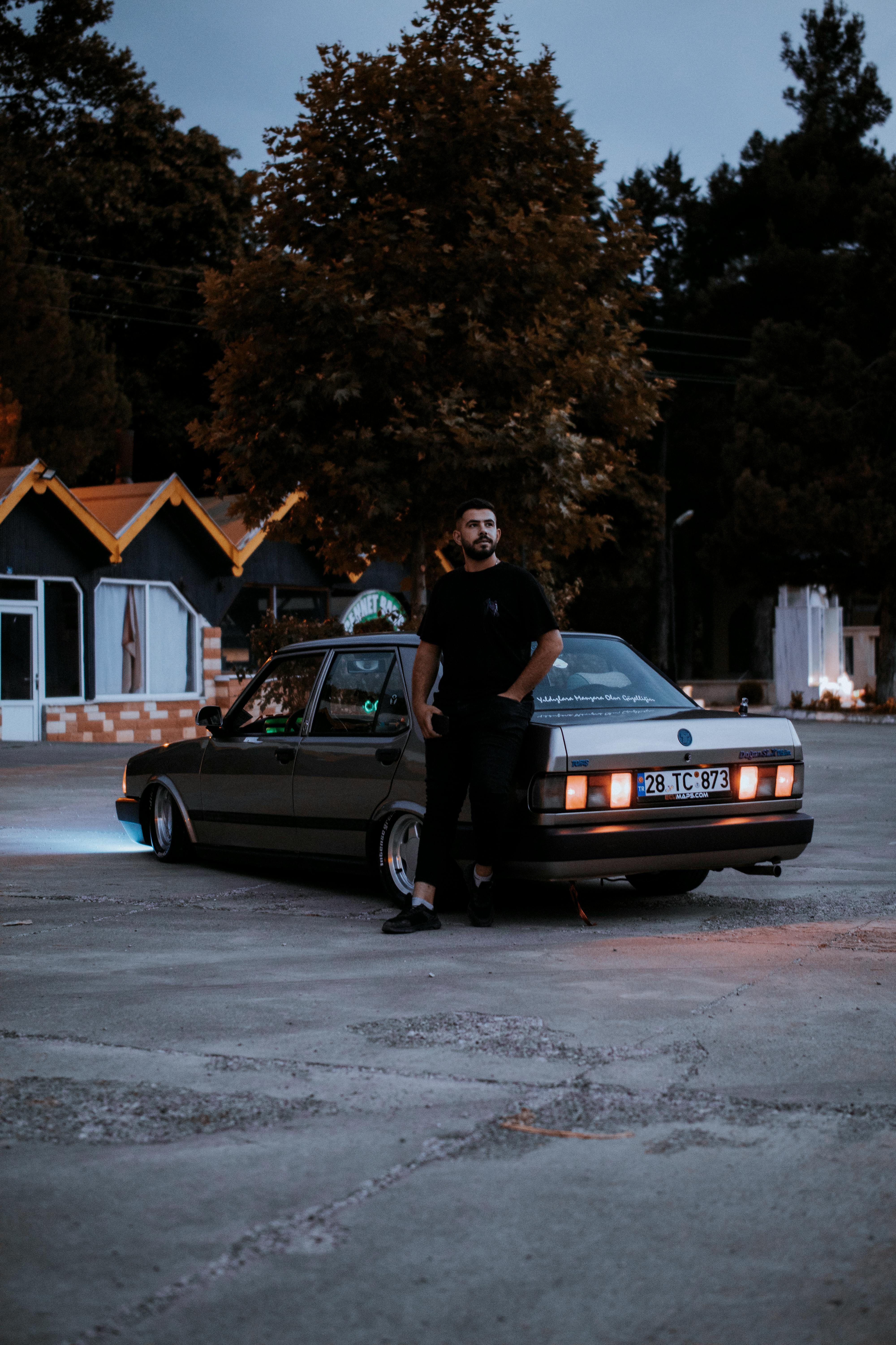 A man standing next to a modified classic Turkish car in a dimly lit parking lot during evening hours.