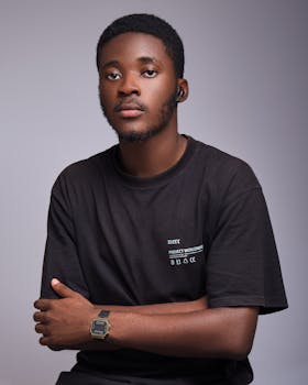 A young Black man with a short hair, wearing a black t-shirt, poses with his arms crossed against a gray background.