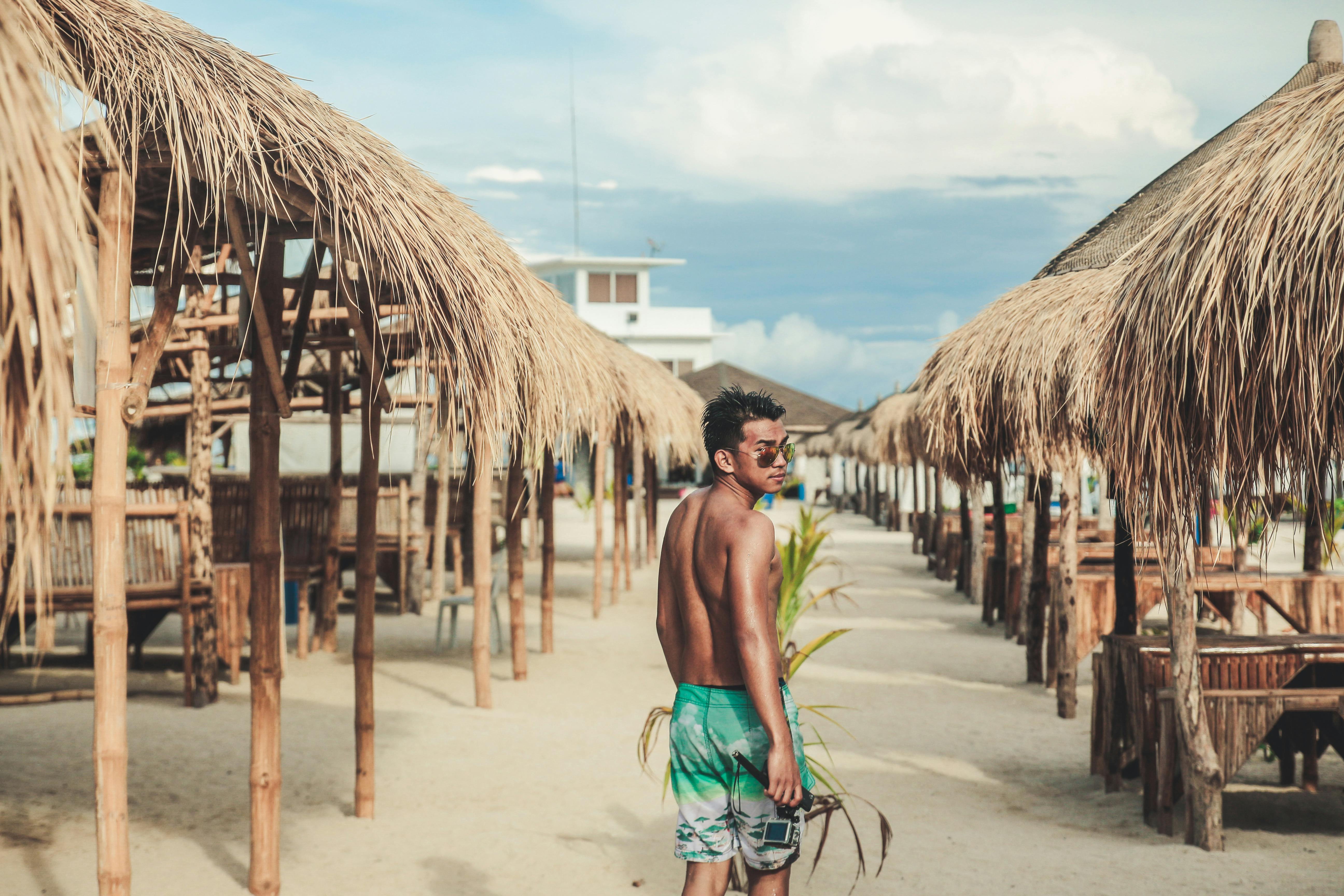 Free Asian man with sunglasses stands at a tropical beach resort under rustic sunshades, enjoying a summer vacation. Stock Photo