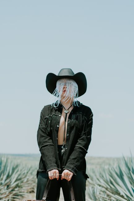 Unrecognizable person posing in a cowboy hat and jacket in a field under a clear sky.