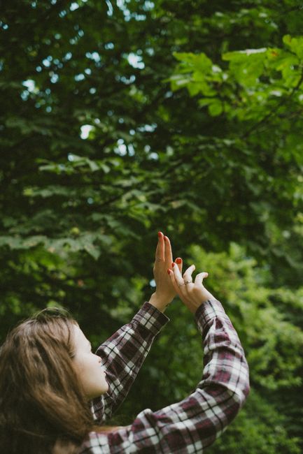 A young woman in flannel reaches up towards lush green leaves in a nature park, creating a serene and contemplative moment.