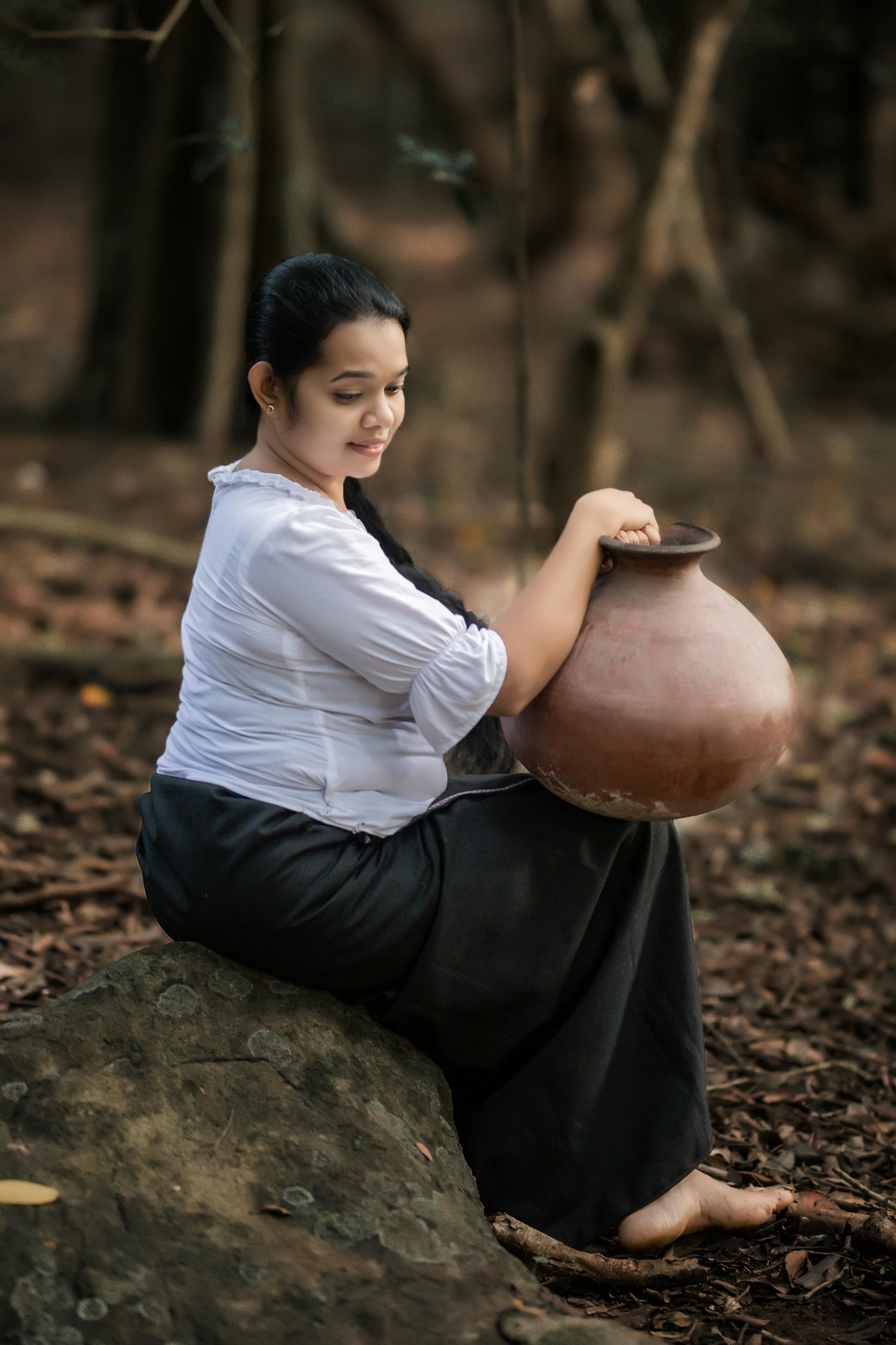 South Asian woman sitting in a tranquil forest setting, holding a traditional clay pot.