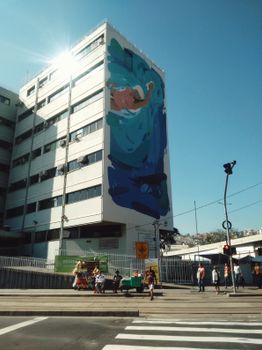 城市街景充满活力，建筑物上有一幅大型壁画，人行道上有行人往来