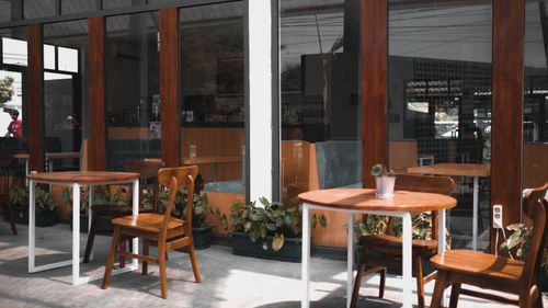 Free Wooden chairs and tables outside a modern cafe with large windows and plant decor. Stock Photo
