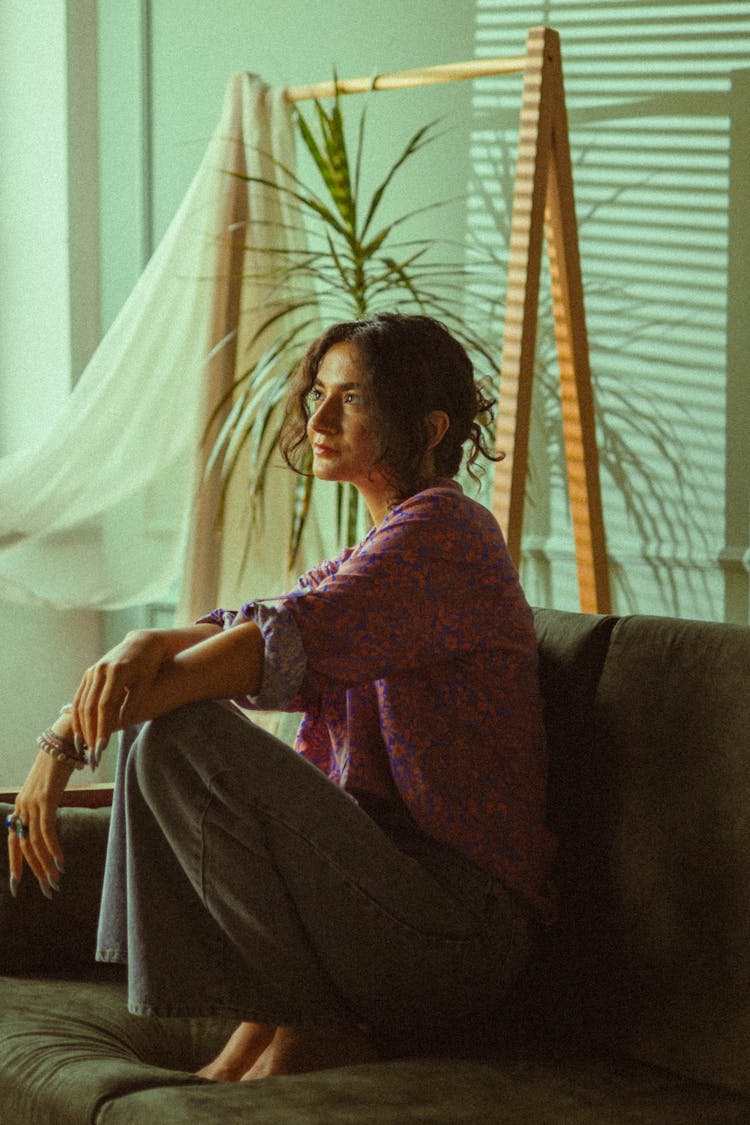 Young Woman In Floral Pattern Blouse And Flare Jeans Sitting On Sofa