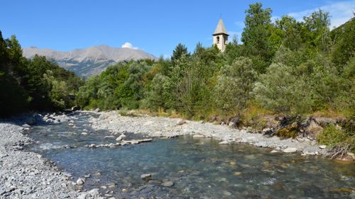 Free A serene mountain river flowing alongside a church tower surrounded by lush forest. Stock Photo