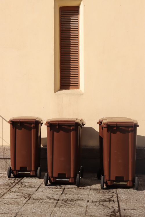 Free Three brown garbage bins lined up against a yellow wall in Alghero, Sardinia, Italy. Stock Photo