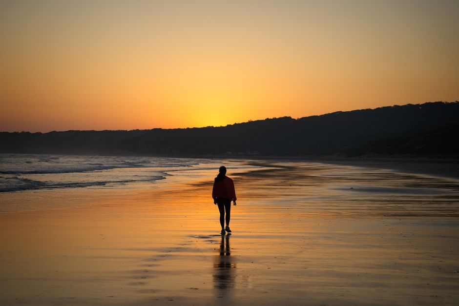 A serene scene of a woman walking along a Melbourne beach during a golden sunset, reflecting on the calm ocean.