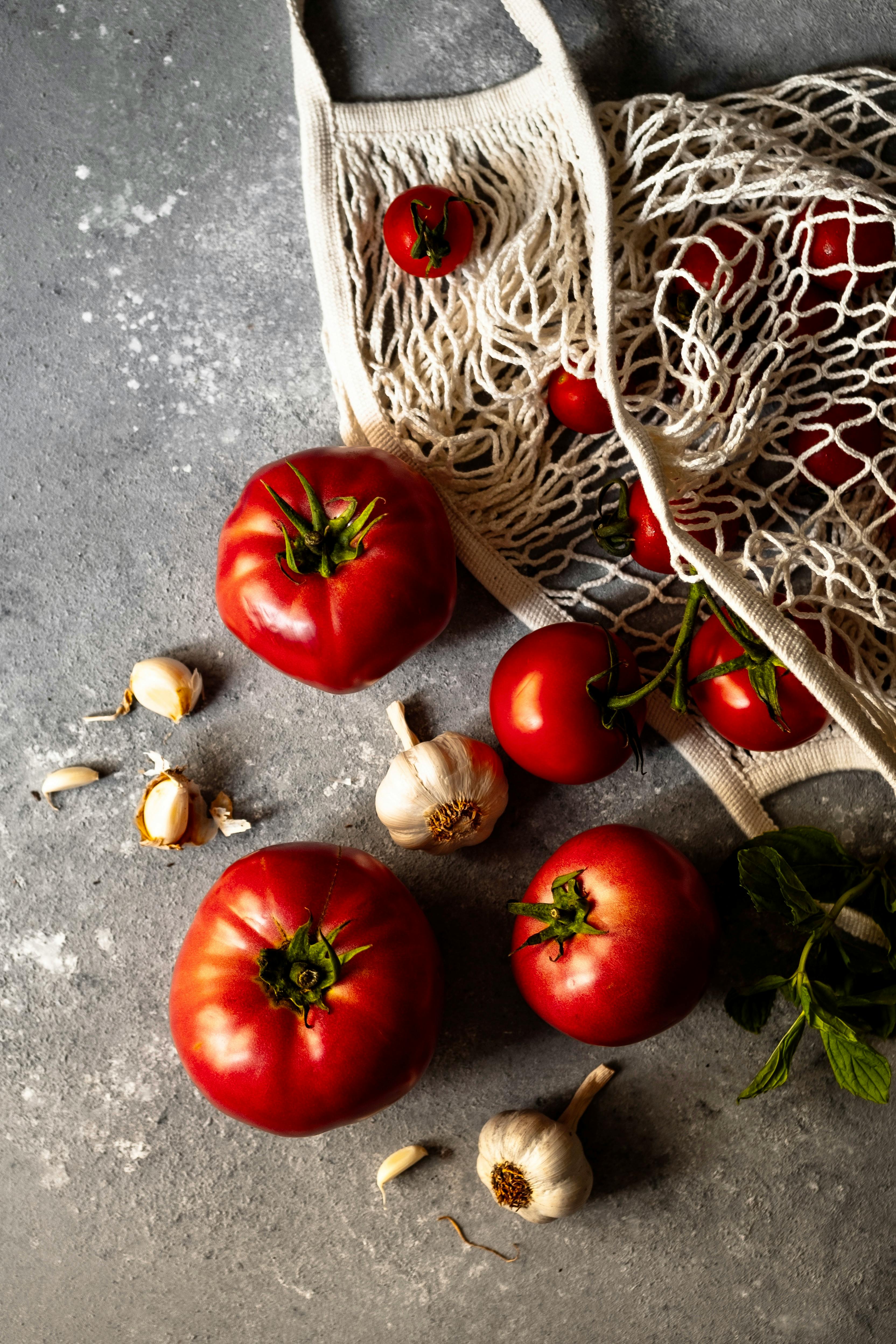 Top view of fresh tomatoes and garlic spilling from a net bag, with a gray background.