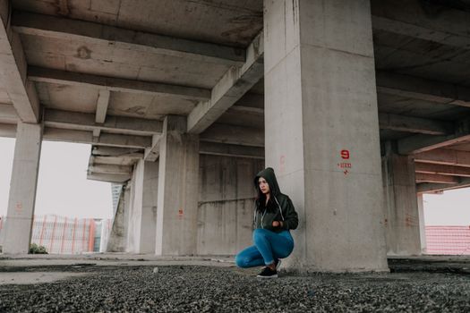 一名身穿连帽衫和牛仔裤的女子在混凝土桥下摆姿势，展现都市时尚