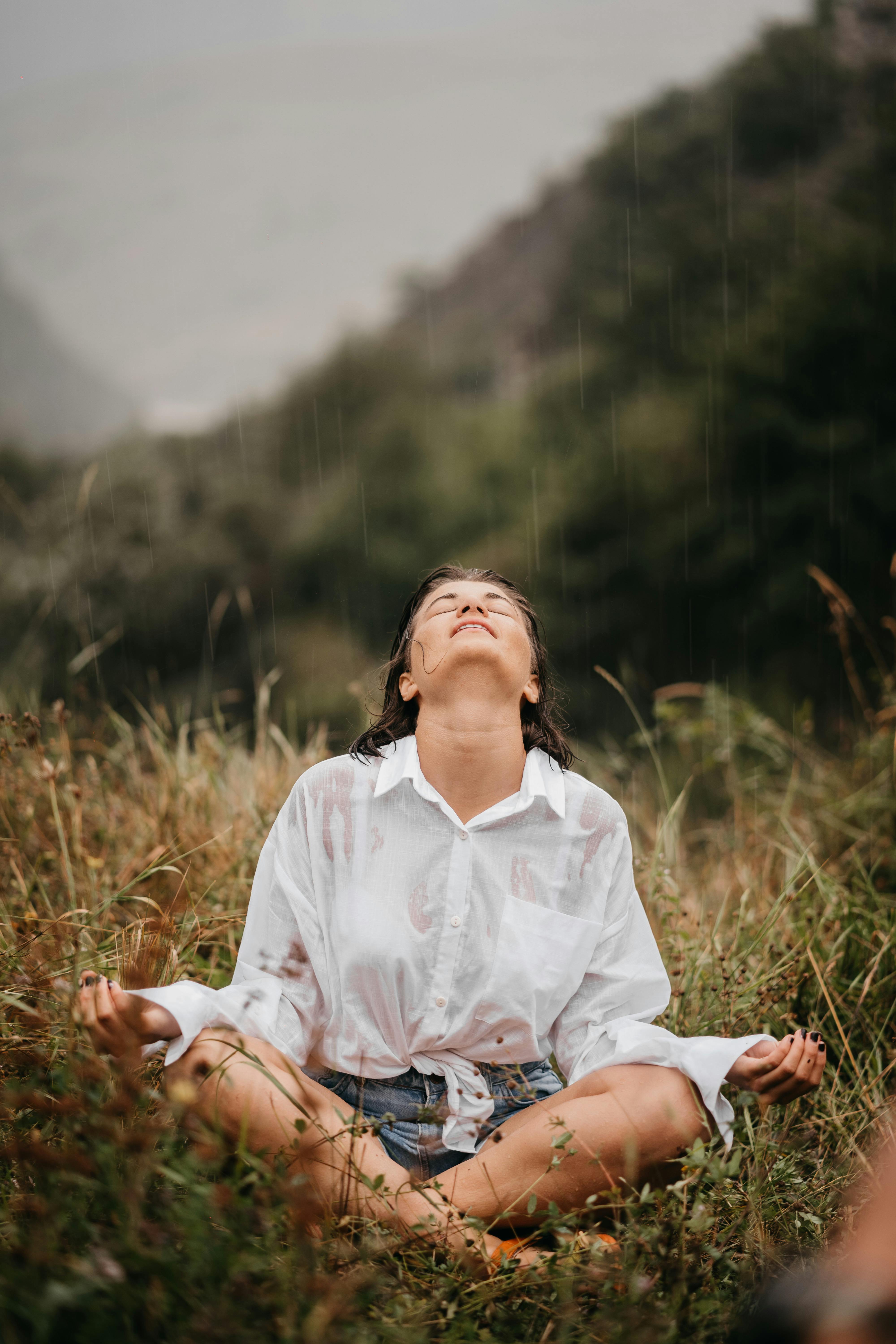 A woman sits peacefully in the rain, embracing nature and mindfulness in a lush outdoor setting.