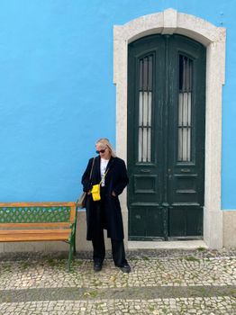 Woman in black coat stands by a blue wall and green door in Lisbon street.