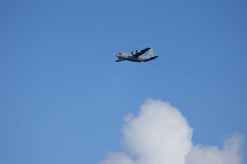 Free Lockheed C-130 Hercules aircraft flying against a clear blue sky with clouds. Stock Photo