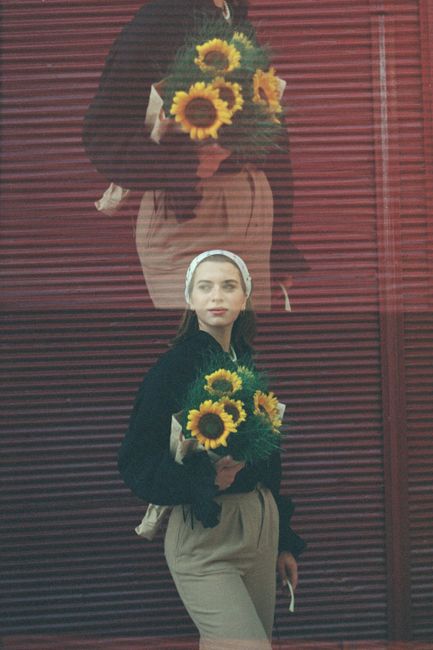 A woman with a sunflower bouquet stands against a reflective city street backdrop.