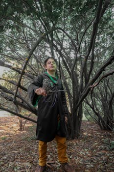 A man in a cloak in a forest holding a bird. Captured outdoors with trees around.