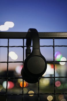 Black wireless headphones hanging on a fence in an urban setting with blurred city lights at dusk.
