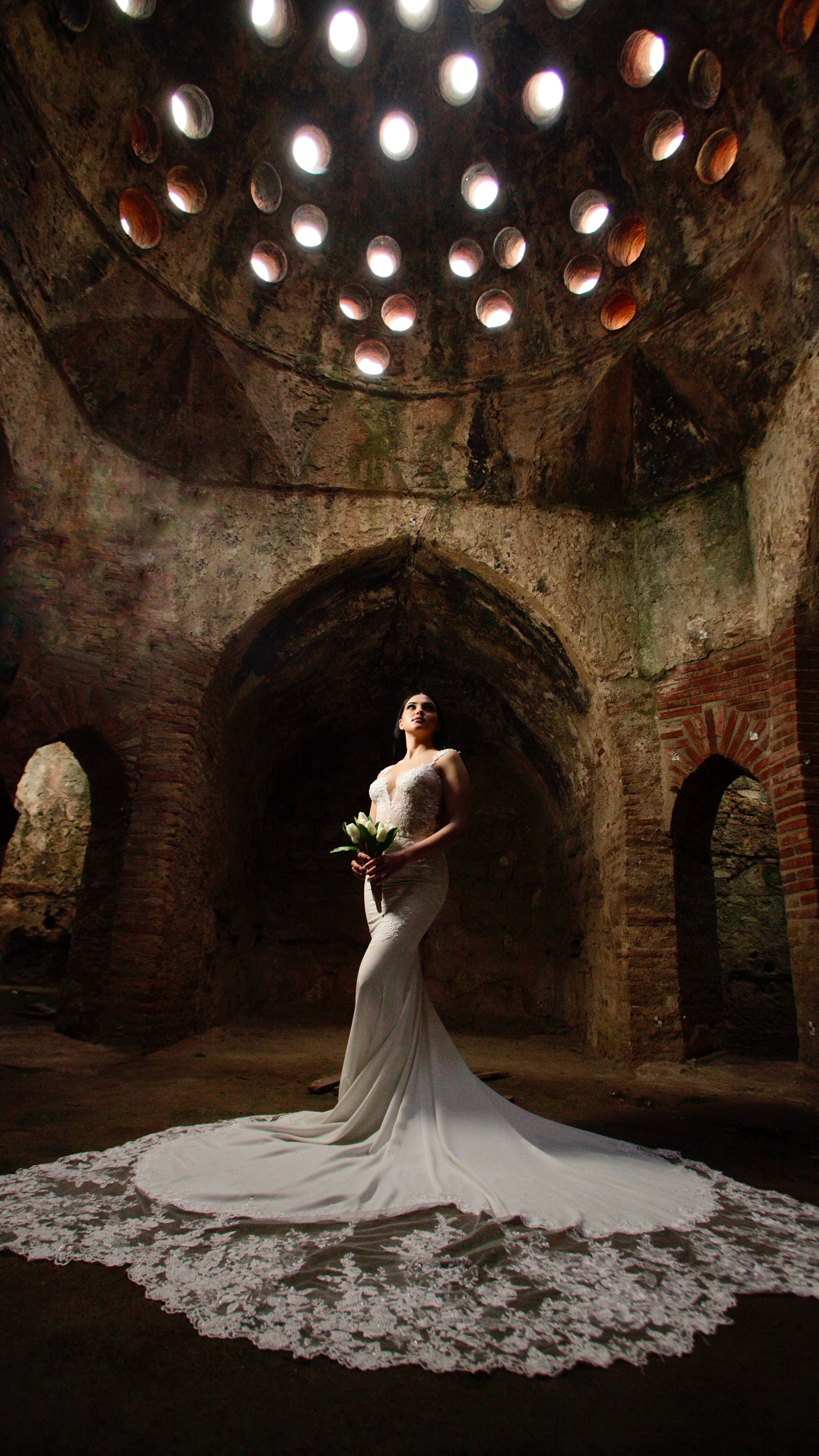 A bride in a white dress holds a bouquet inside a historic stone chamber with unique lighting.