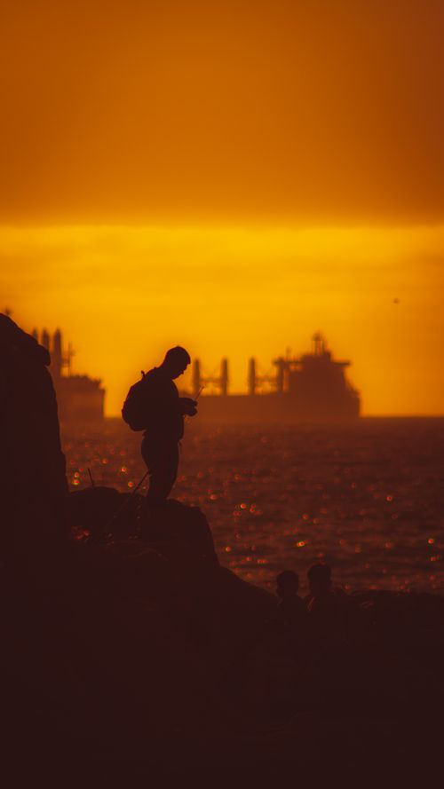 Free A silhouette of a person on a rocky shore against a vibrant sunset in Viña del Mar, Chile, with ships in the sea. Stock Photo