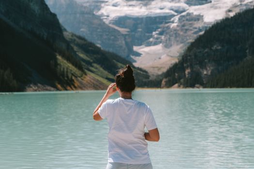 A woman in a white t-shirt stands by a mountain lake, enjoying the serene view.