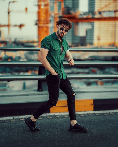 Free Stylish man with sunglasses in green shirt and ripped jeans posing on bridge against urban backdrop. Stock Photo