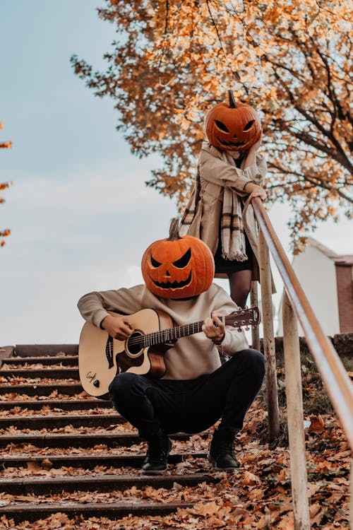 Man Playing Guitar and a Woman Walking Down the Stairs Littered with Autumn Leaves Both with Carved Pumpkins on Their Heads