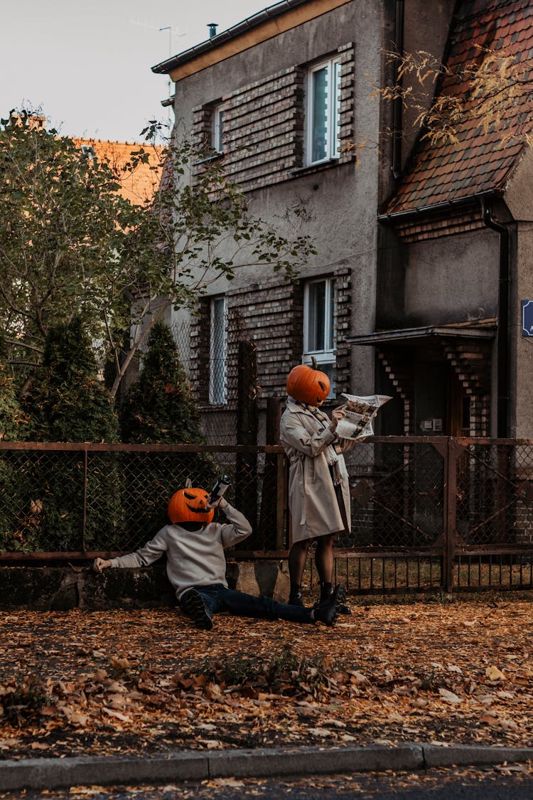 People With Pumpkin Heads Standing And Sitting Near House In Autumn