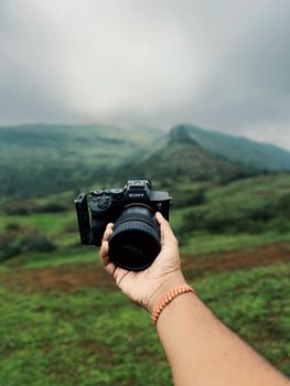 A hand holding a camera with lush green Indian mountains in the background, capturing nature's beauty.