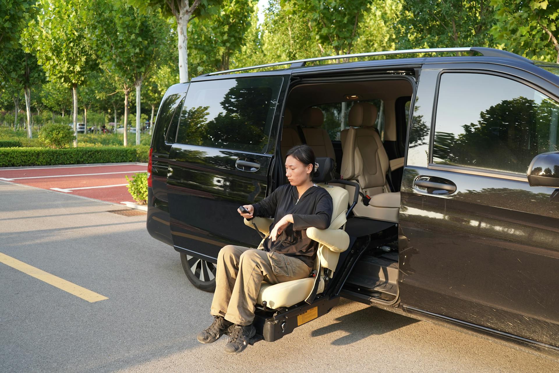 Woman using a seat lift in a black van at a parking lot with trees in the background.