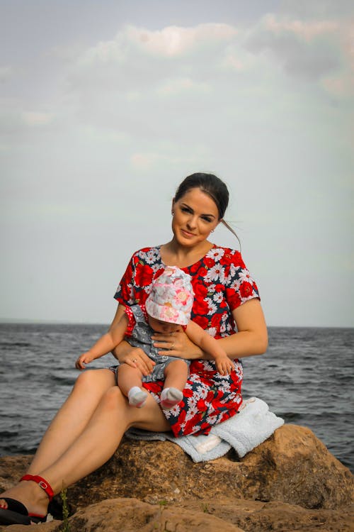 Smiling Mother in Sundress Sitting with Baby on Rock on Shore