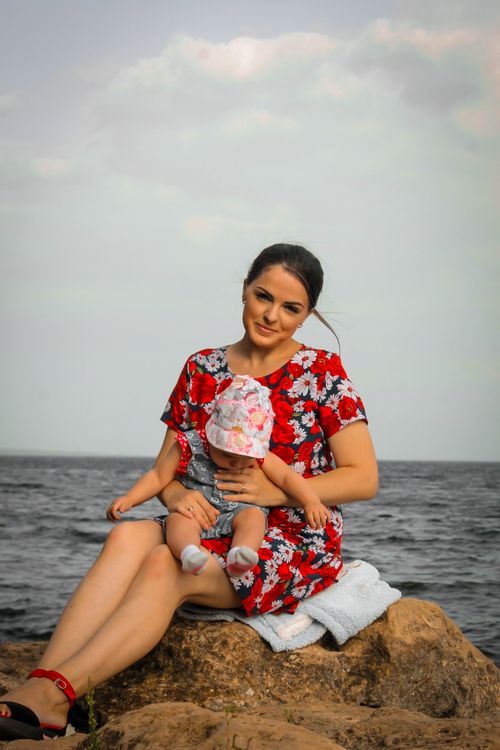 Free Woman in floral dress holds baby while sitting on rocks by the sea. Stock Photo
