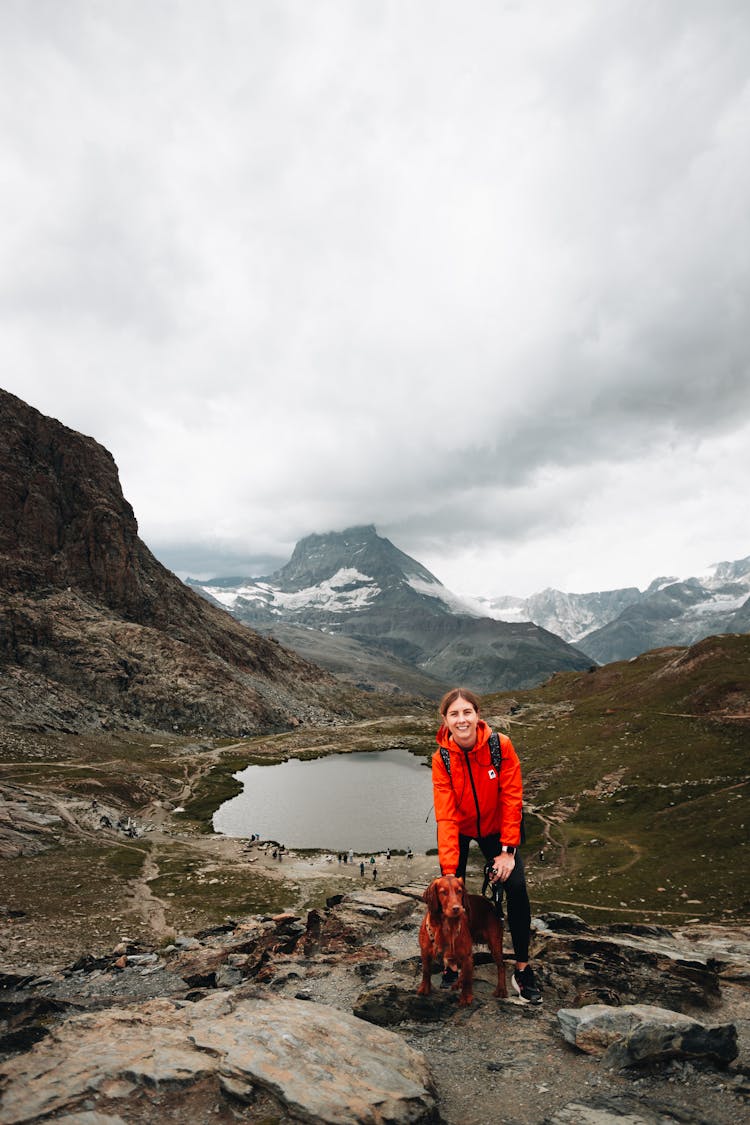 Woman In Red Coat Standing On Mountain Trail Overlooking Lake With Dog