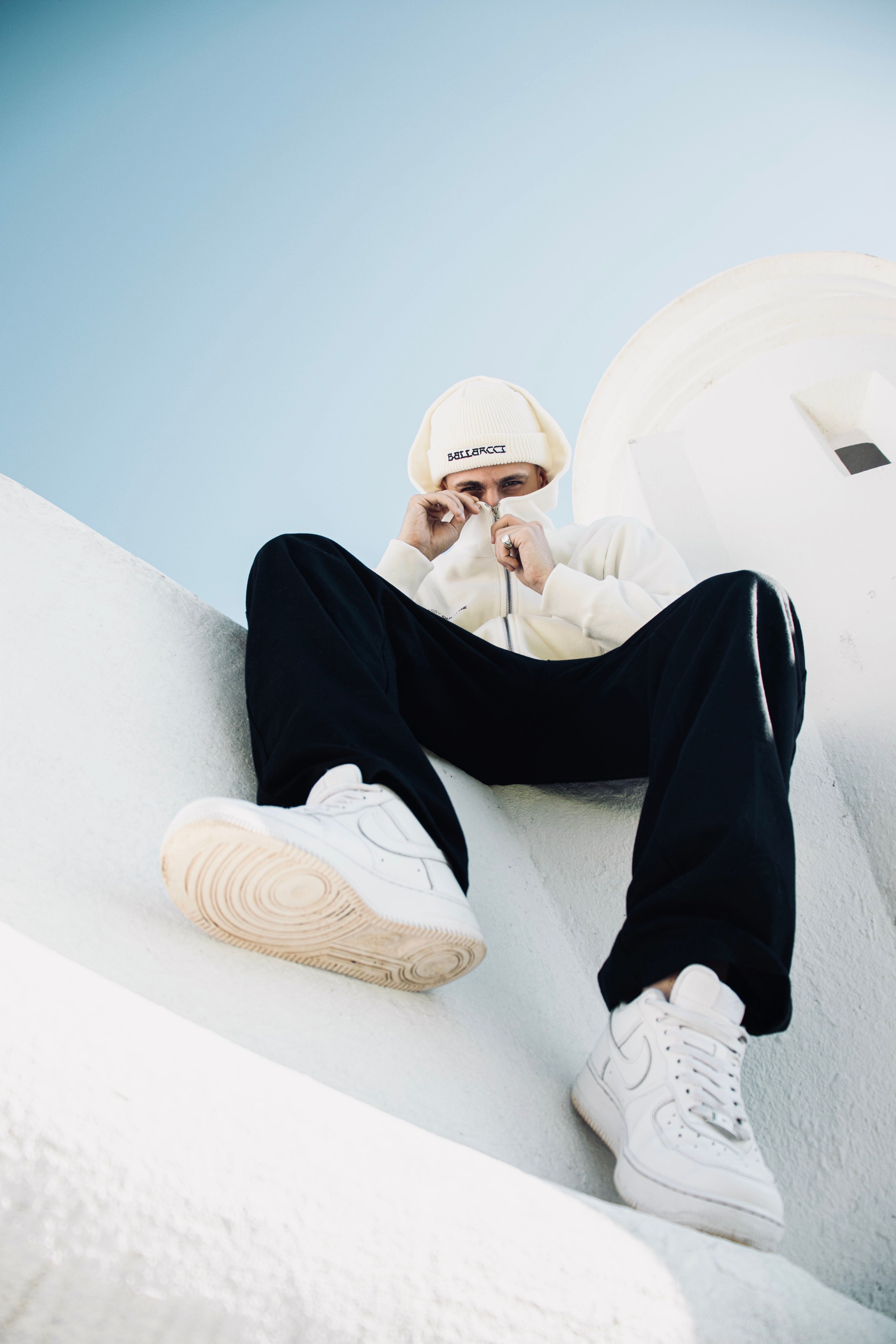 Low-angle shot of a man in urban attire posing against a white wall, showcasing street style.