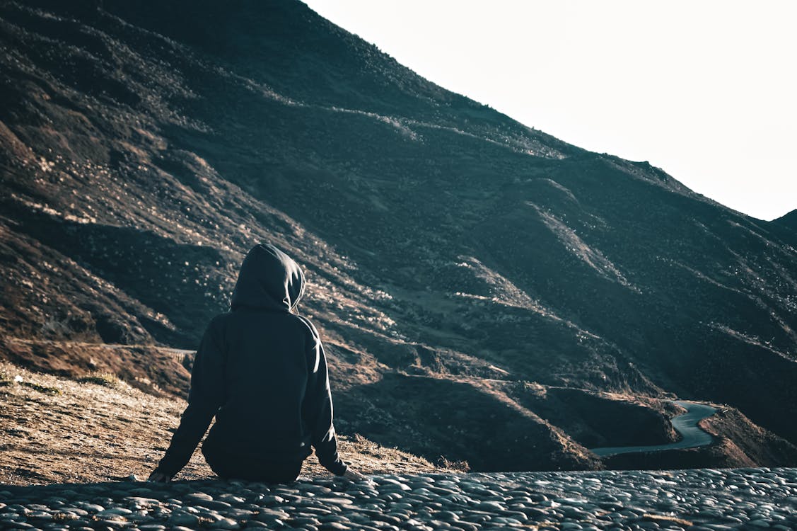 Person in Hooded Jumper Sitting on Ground above Mountain Valley