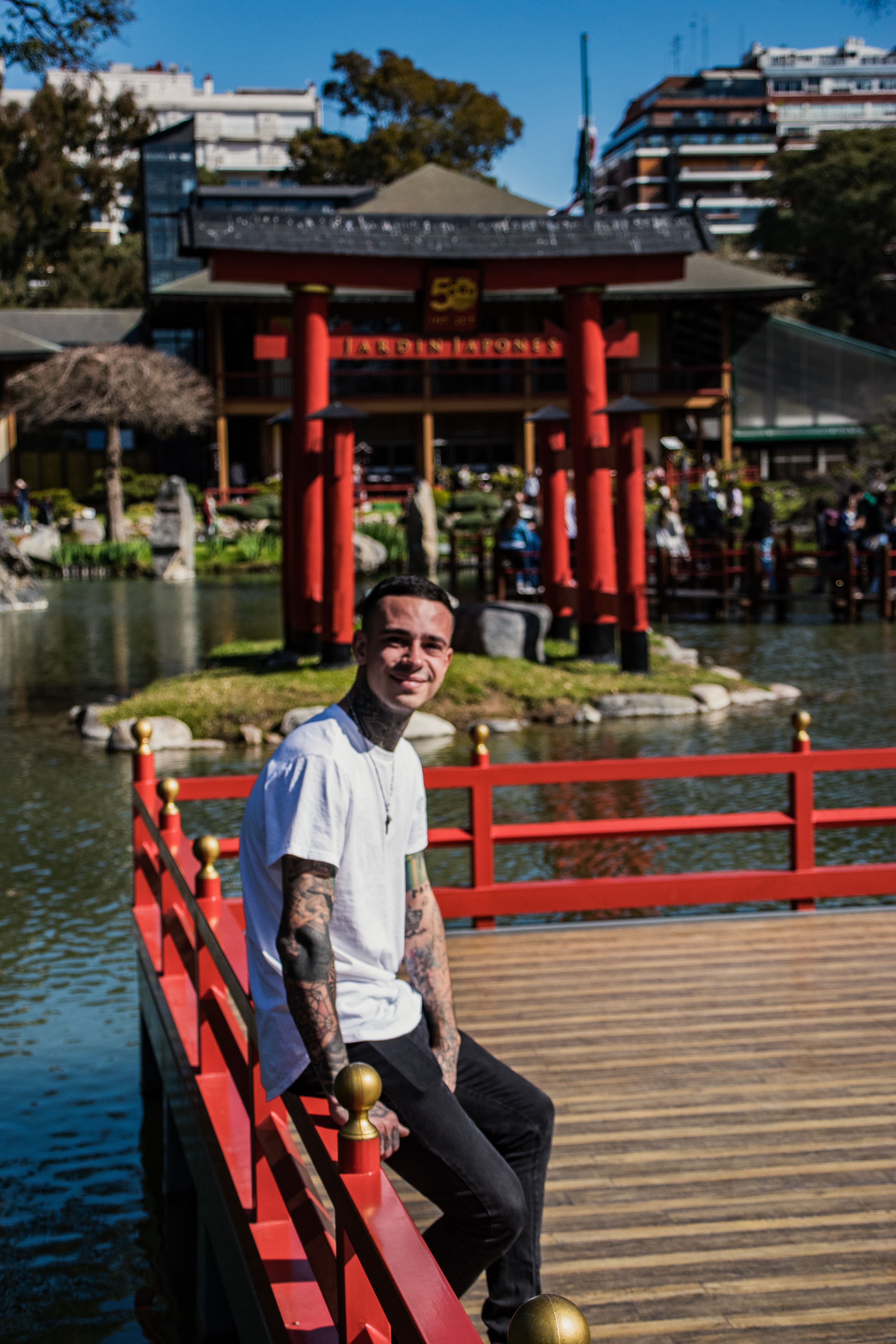 Tattooed man sitting on rail by pond in Japanese garden with red structures.