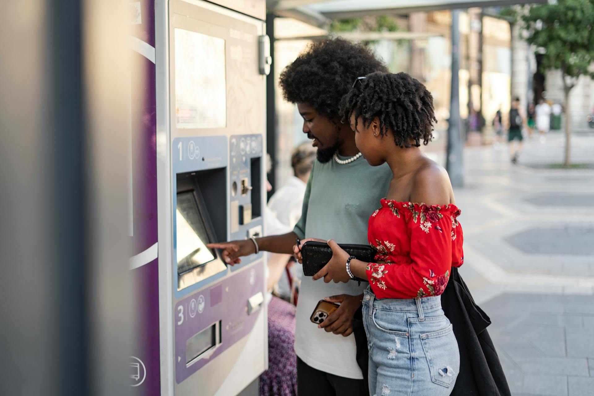 A young couple buying tickets using a touchscreen machine at an urban bus stop on a sunny day.