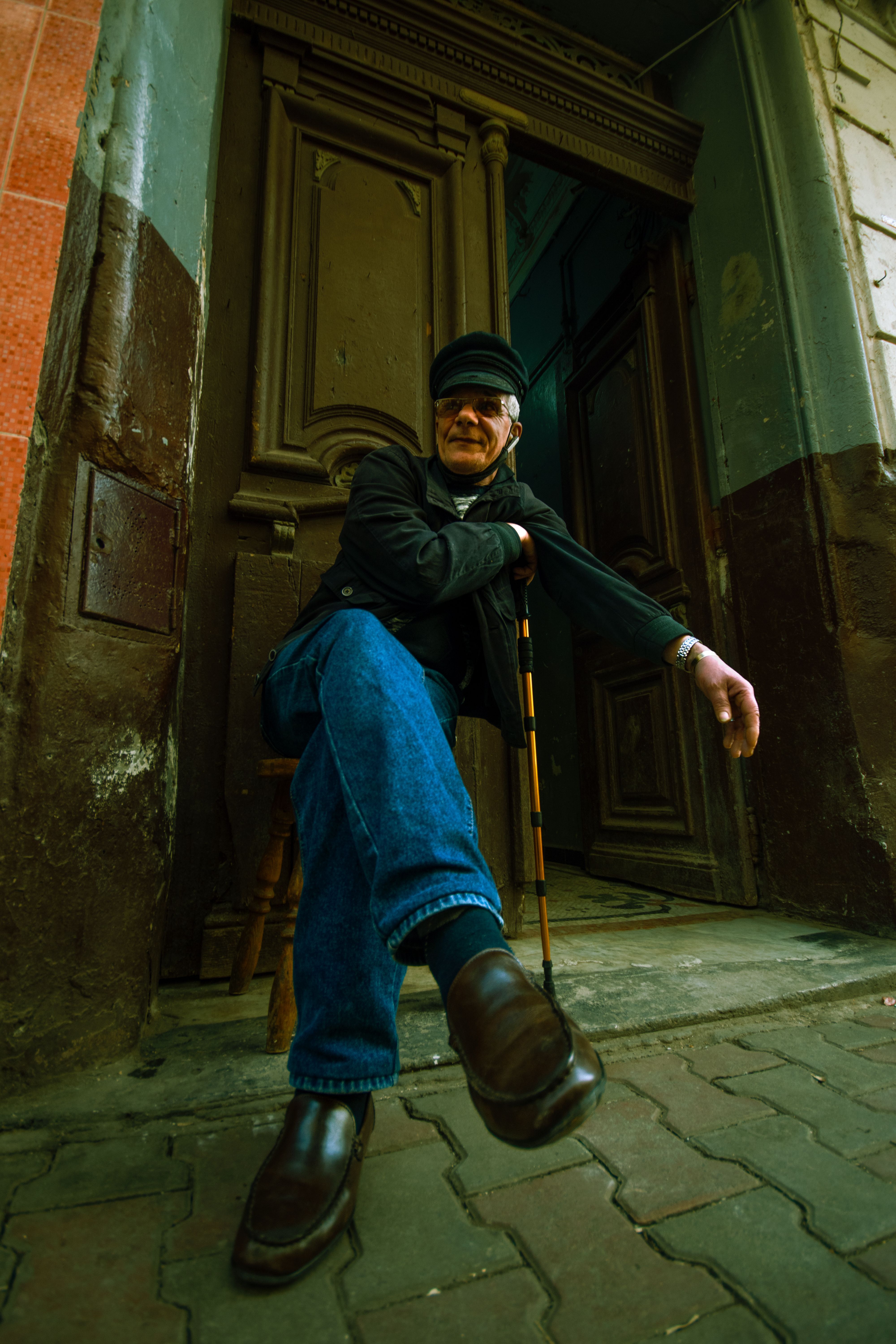 Elderly man sitting with cane in front of a wooden doorway on a charming city street.