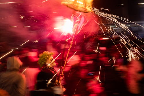 Group of People Blurred with a Person in a Costume Holding a Stick with Small Colourful Fireworks
