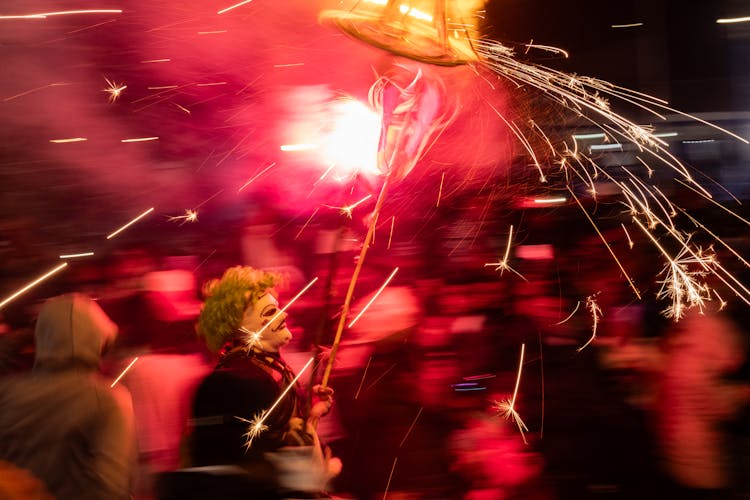 Group Of People Blurred With A Person In A Costume Holding A Stick With Small Colourful Fireworks