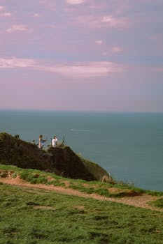 Three people relax on a grassy cliff overlooking the sea during sunset.
