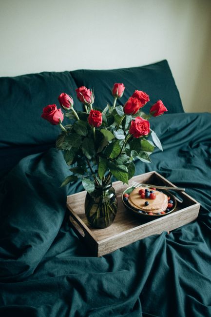 A cozy breakfast in bed setting featuring red roses and pancakes with berries on a wooden tray.