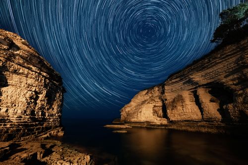 Free A mesmerizing long exposure of star trails above rugged rocky cliffs by the sea at night. Stock Photo