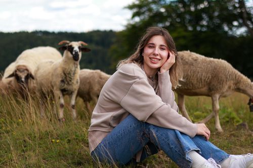 Free A young woman sitting in a rural pasture, smiling among sheep. Stock Photo