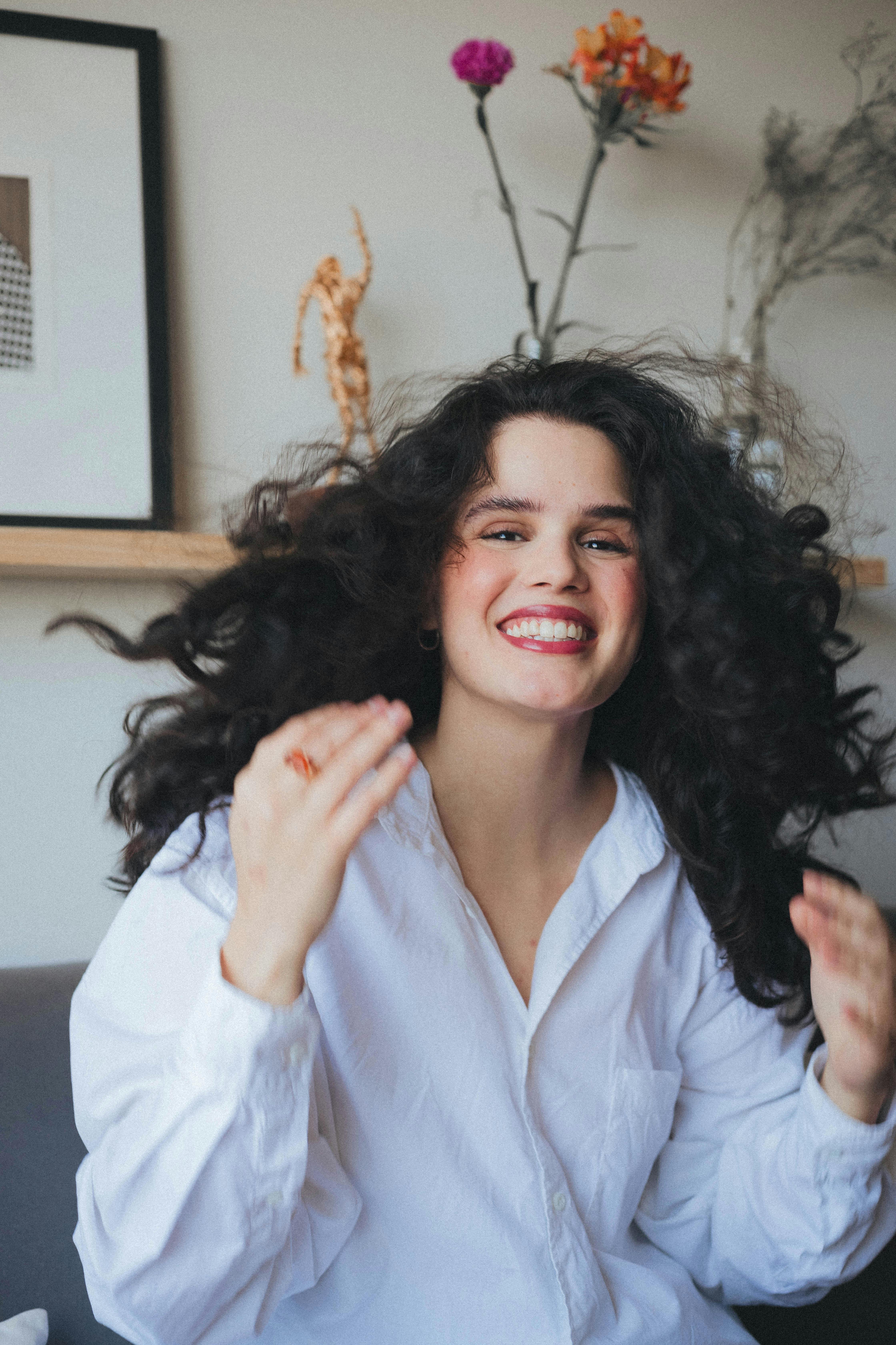 Portrait of a smiling woman with long black hair wearing a white shirt in a cozy indoor setting.