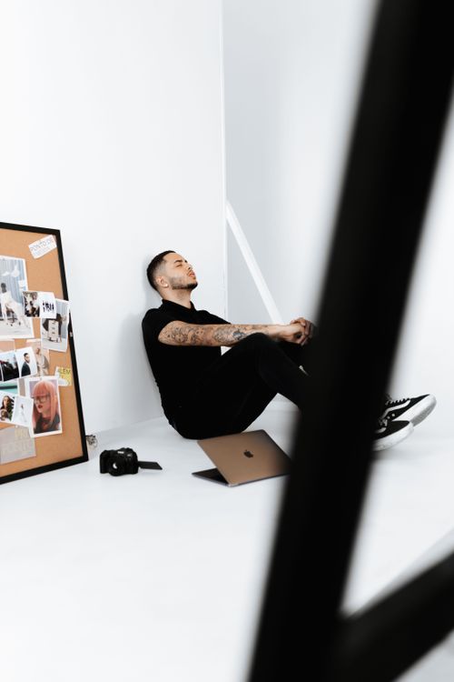 Free A man sits against a white wall in a minimalist workspace with a laptop and camera. Stock Photo